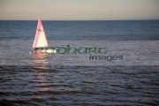 small-pink-white-topaz-dinghy-sail-boat-sailing-across-the-water-in-evening-sunlight-reflecting-in-the-water