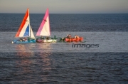 small-pink-white-red-white-blue-topaz-dinghy-sail-boats-sailing-being-towed-by-inflatable-across-the-water-in-evening-sunlight